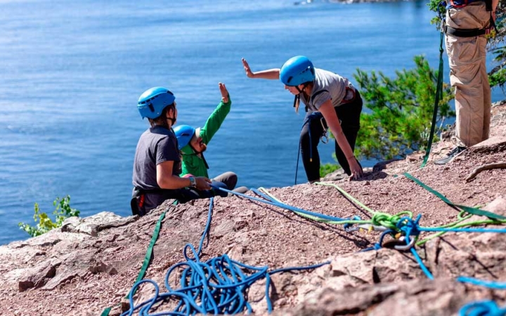 two people who are rock climbing high five just over the edge of a cliff. There is blue water below them.